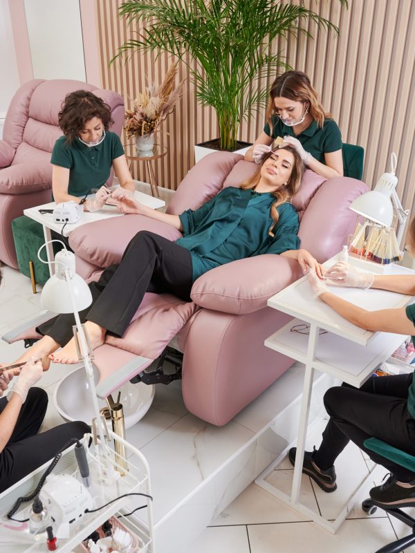 Manicurist, pedicurist and eyelash specialist working with client in modern beauty salon. Woman relaxing in comfortable salon chair while having manicure, pedicure and eyelash extension procedure.